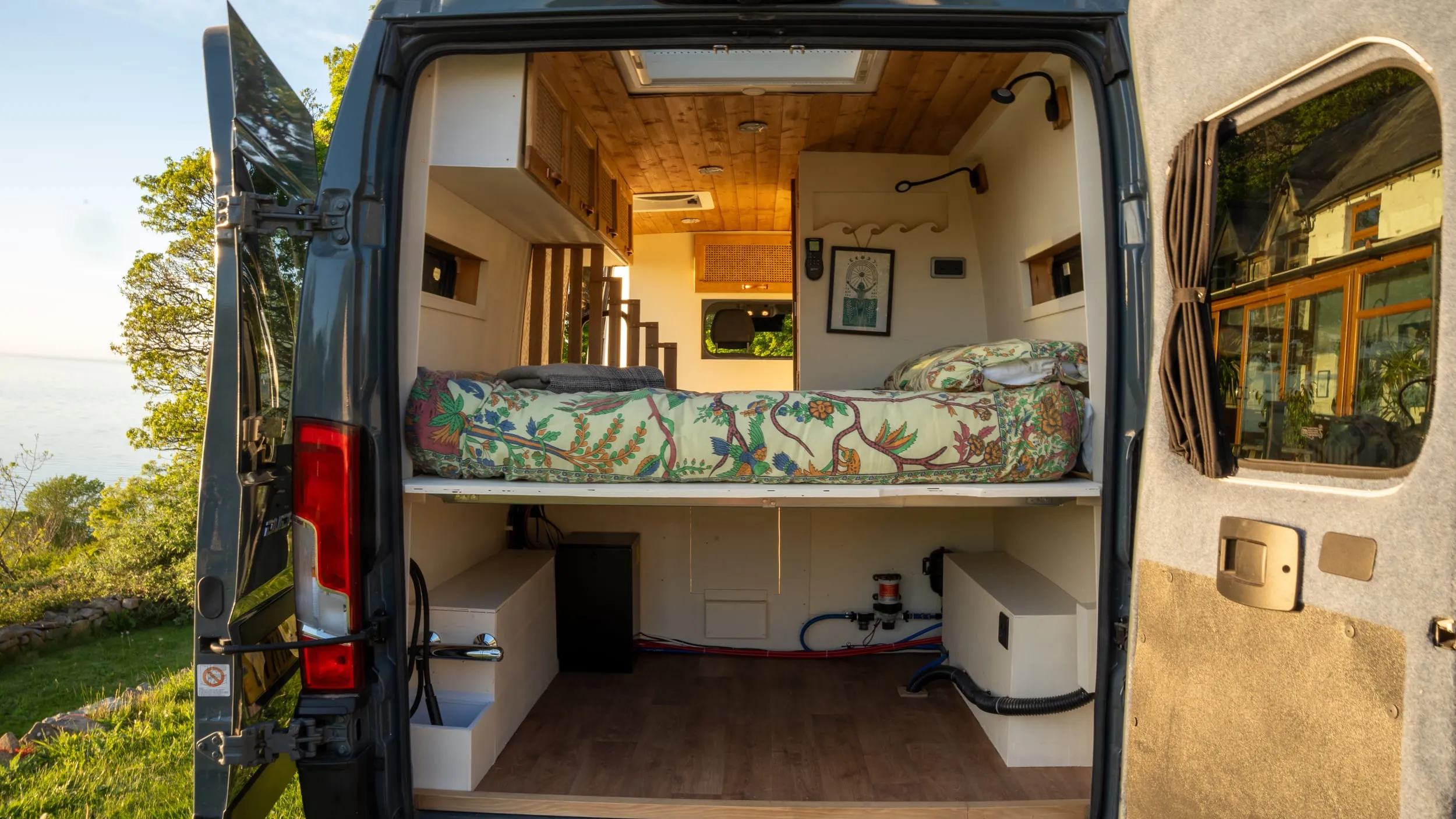The finished van through the open rear doors: bed across the back, galley down one side, timber-lined ceiling.