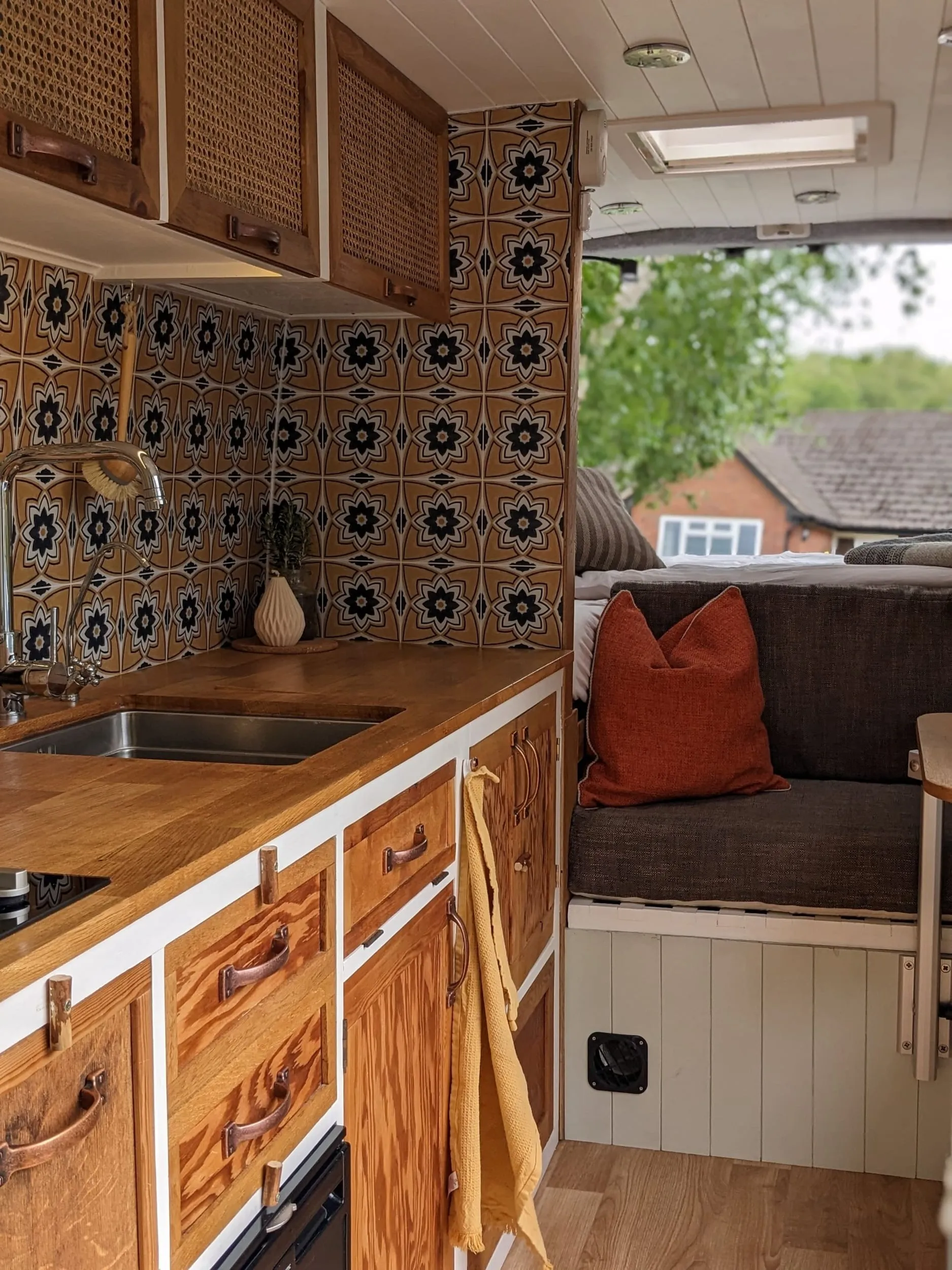 Inside a finished campervan: an oak worktop with a sink and tap against a patterned tiled splashback, cane-fronted lockers above, reclaimed timber drawers with copper handles below, and round to a bench seat with a rust cushion. A roof light above, and the diesel heater outlet vent set into the panelling near the floor.