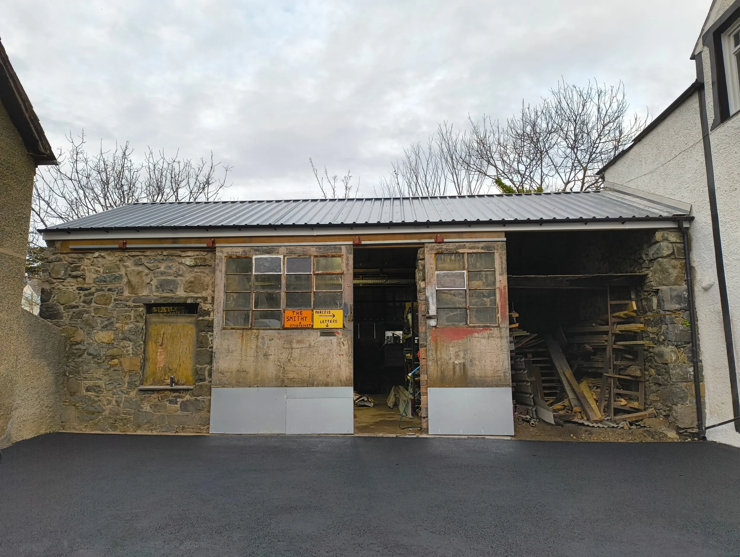 The Smithy, my workshop in Llanfairfechan: an old stone building with a grey metal roof, big sliding doors with the Smithy sign on them, one standing open, and timber stacked in the open bay.